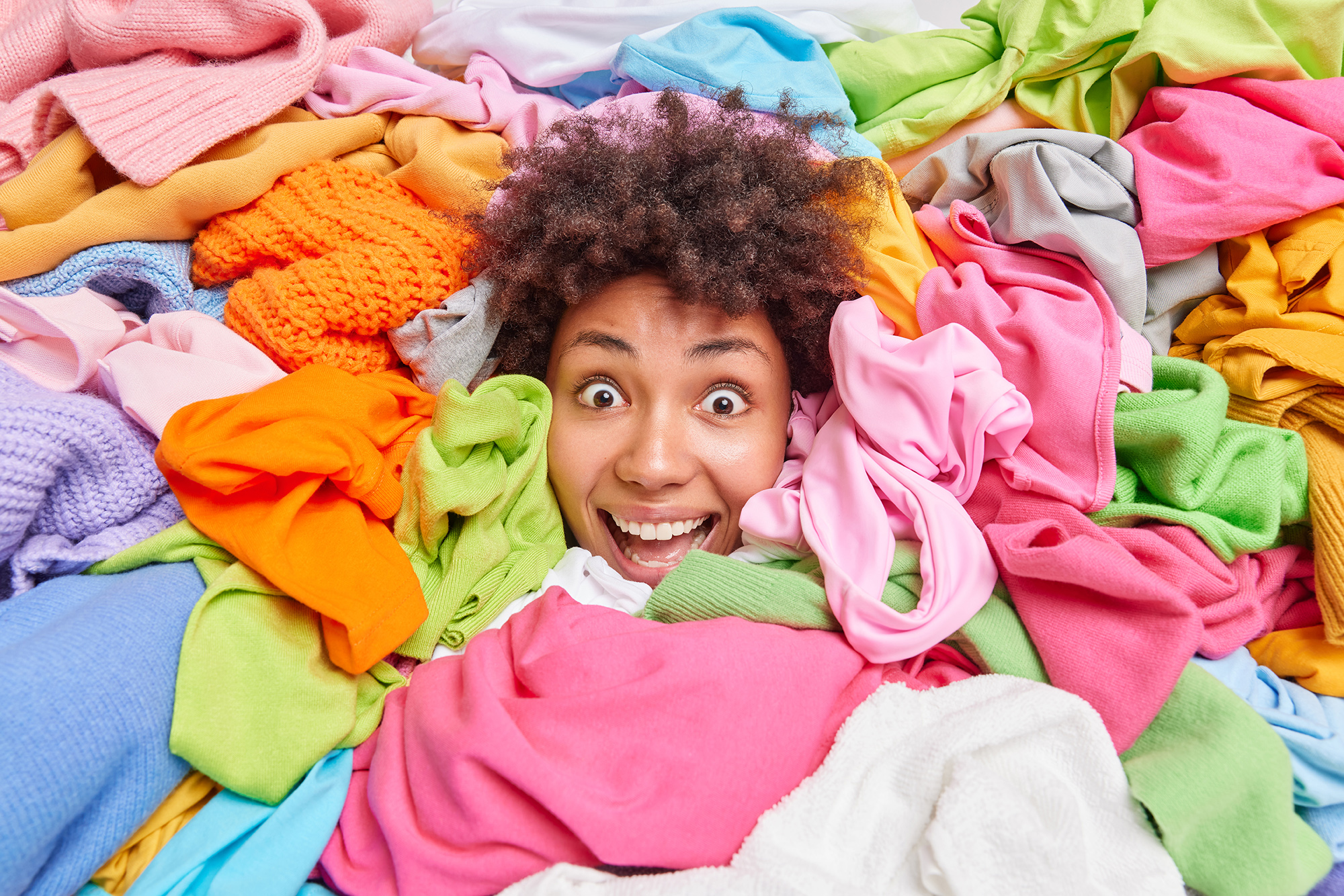 Decluttering and tidying up concept. Impressed positive curly Afro American woman sorts out clothes in wardrobe covered with stack of multicolored clothing organises closet resells used apparels